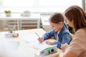 A young girl in a denim jacket focuses on her writing at a table with colorful stationery and notebooks, while seated next to an adult.