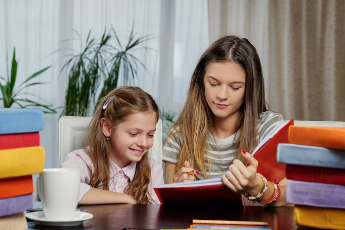 A woman and a girl are engaged in reading a book at a table surrounded by colorful stacked books and a cup of coffee.