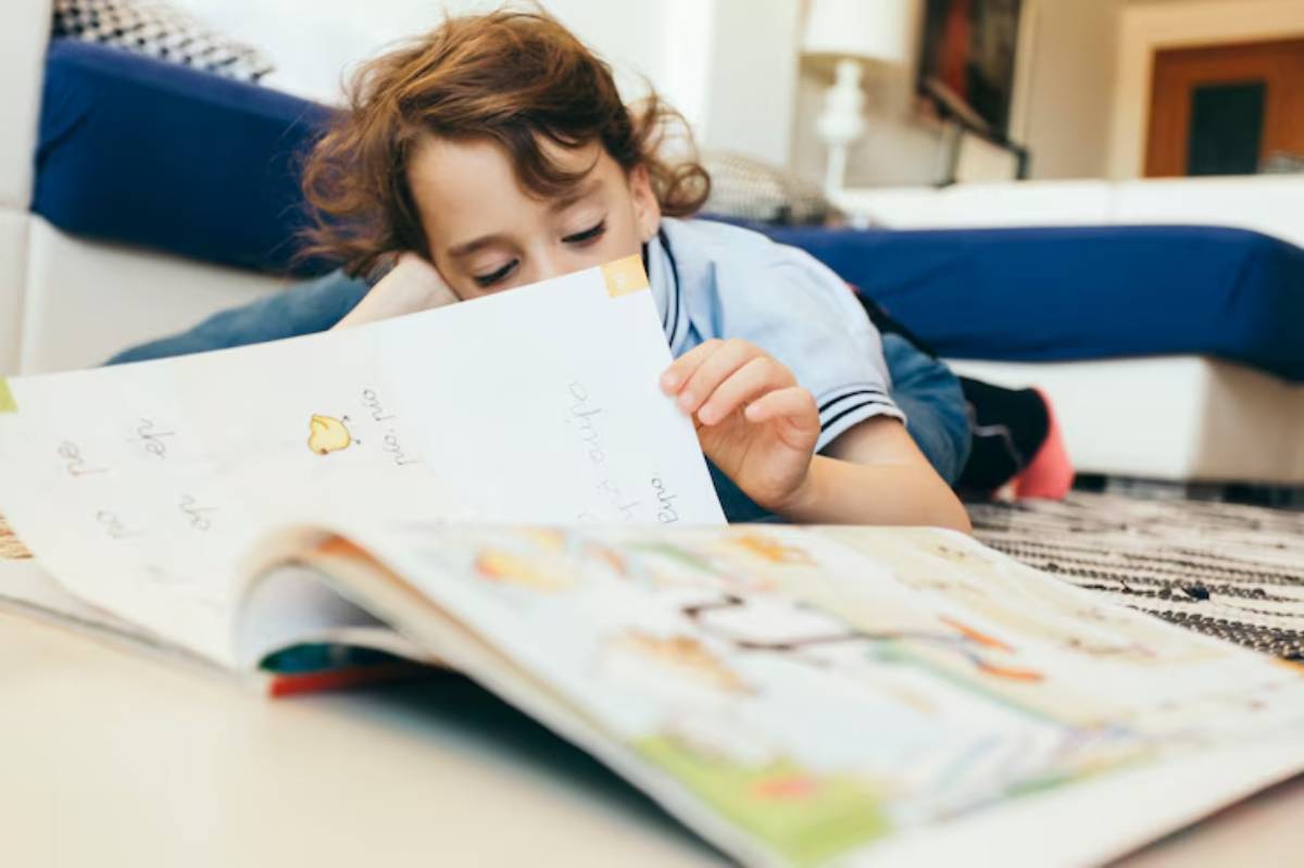 A child rests on their stomach, engrossed in reading a colorful picture book on a patterned rug, with a cozy living room backdrop.