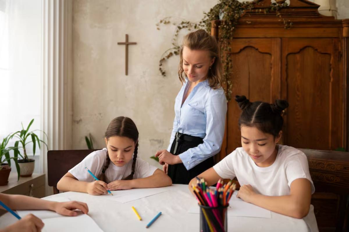 A teacher supervises students drawing at a table filled with colored pencils, surrounded by greenery and a vintage wooden cabinet.
