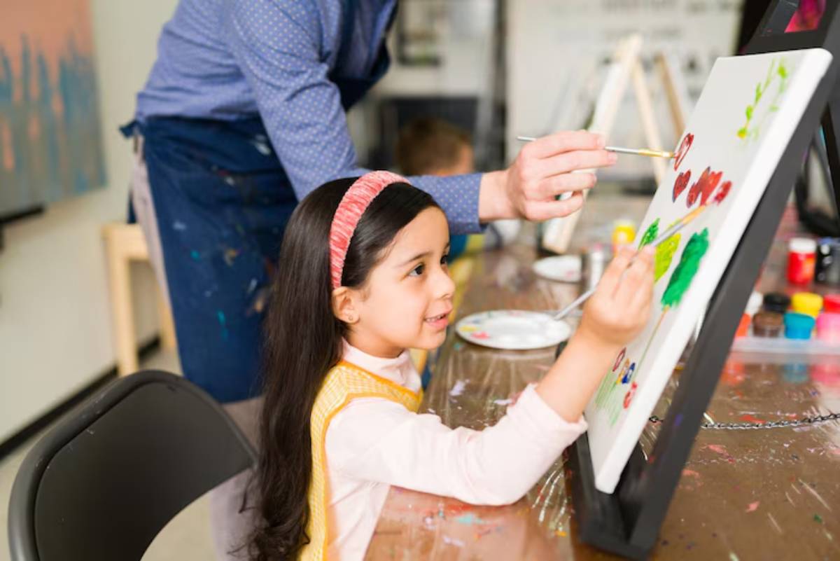 A young child paints on a canvas while an adult guides them, surrounded by colorful art supplies in a bright studio.