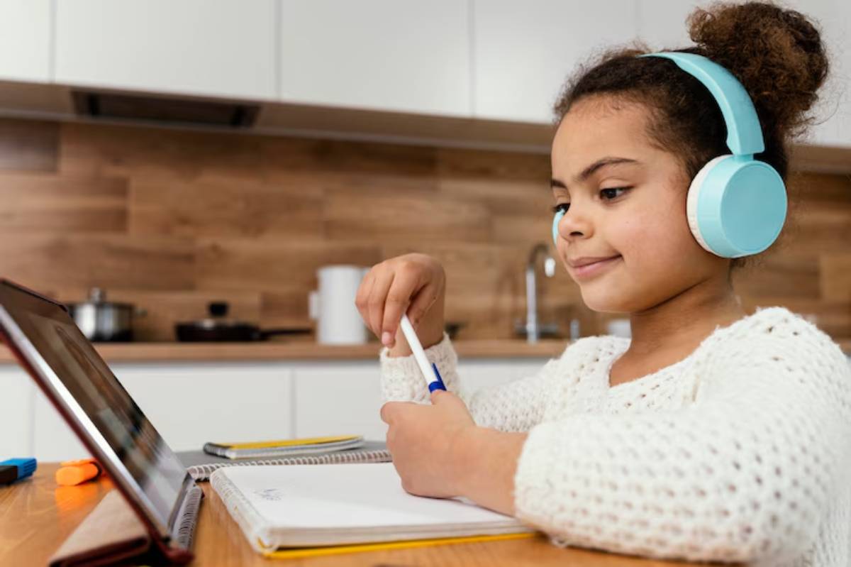 A child wearing blue headphones sits at a kitchen table, focused on a laptop and holding a pen while studying. Notebooks and stationery are visible.