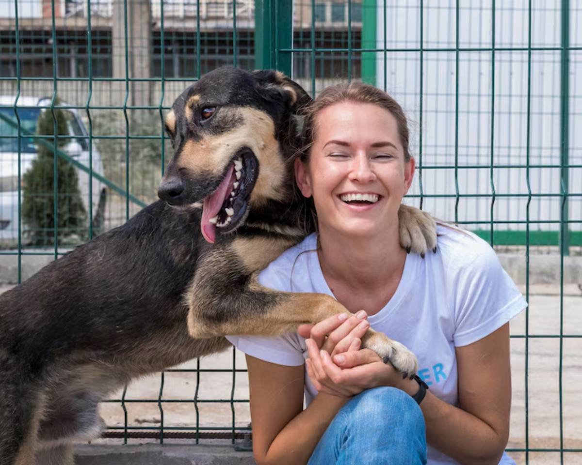 A happy dog rests its front paws on a person's lap, both sitting against a green fence in an outdoor setting.