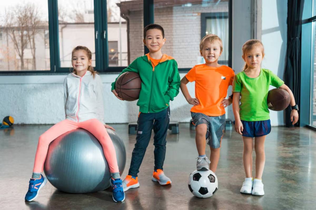 Four children stand in a bright indoor gym, each holding different sports equipment: basketballs and a soccer ball, showcasing active play.