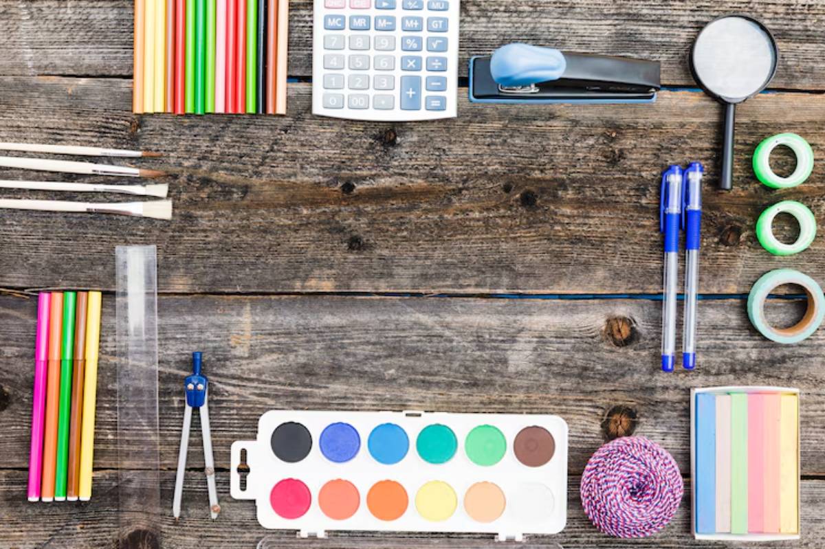 A flat lay of assorted stationery items, including markers, watercolor set, stapler, pens, ruler, compass, and tape on a wooden surface.