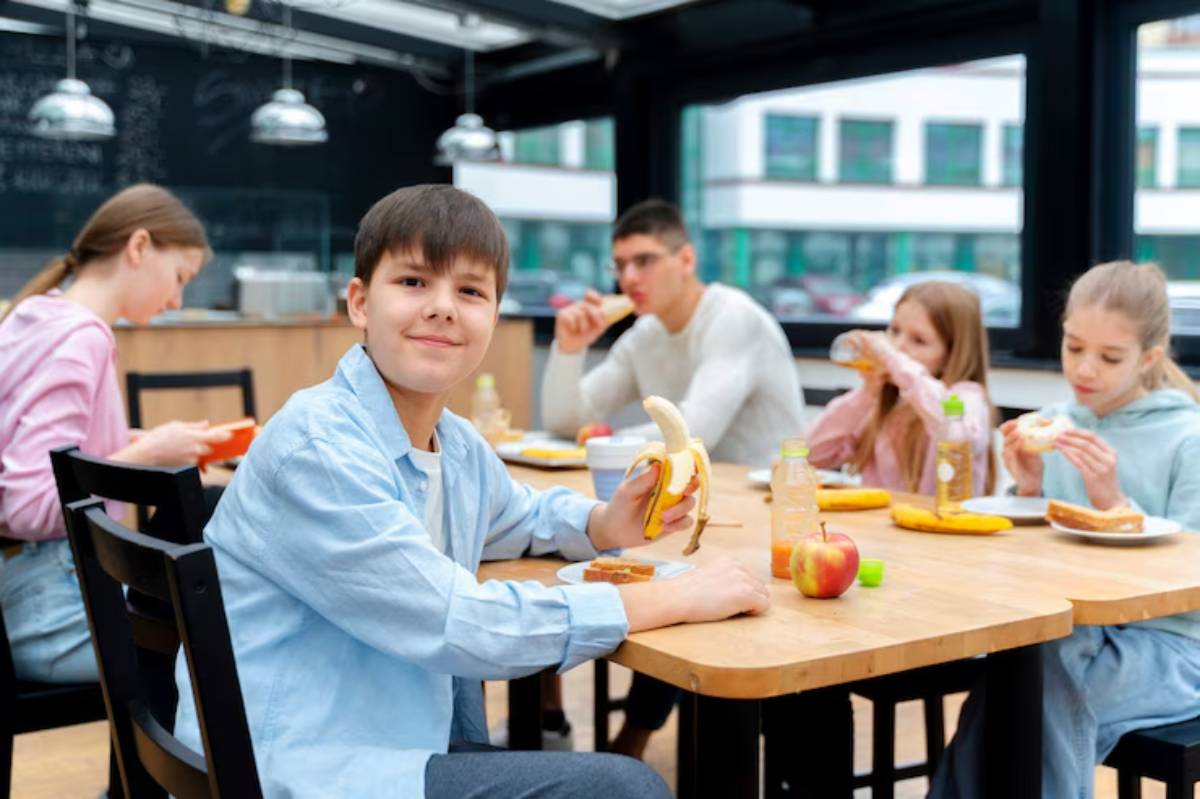A group of young people sits around a dining table, enjoying various snacks, including fruit and sandwiches, in a bright, modern café.