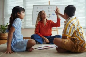 A teacher interacts with two children on a carpet, with colorful educational materials spread out and a whiteboard displaying letters behind them.