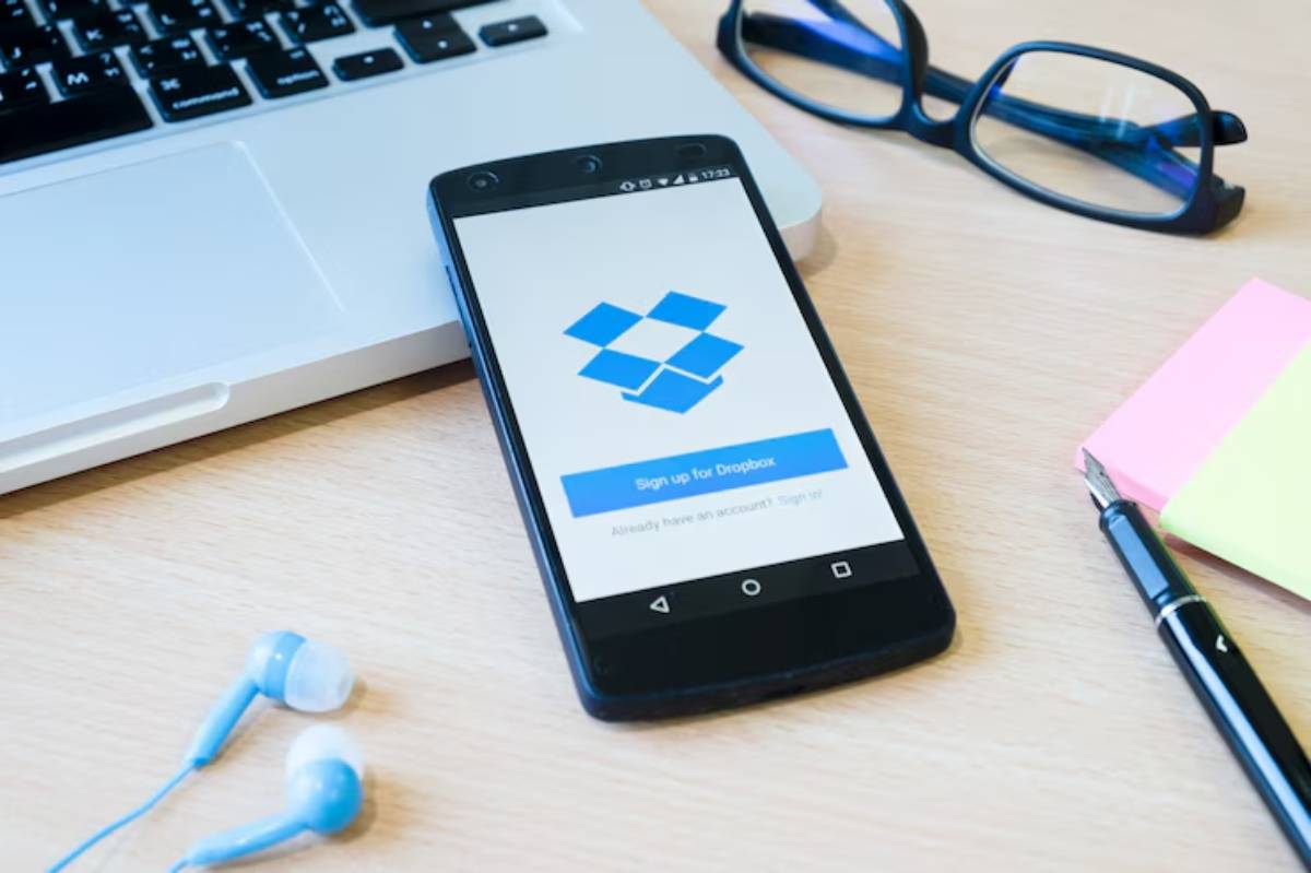 A smartphone displaying the Dropbox sign-up page, placed on a wooden desk with a laptop, glasses, headphones, and sticky notes.
