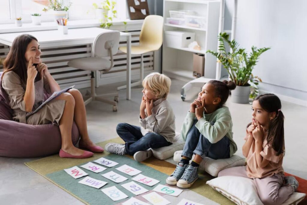 A teacher engages four children sitting on cushions, surrounded by educational materials and a bright classroom setting with plants.
