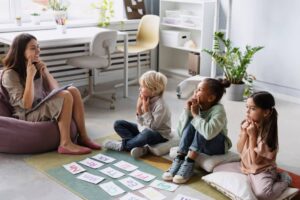 A teacher engages four children sitting on cushions, surrounded by educational materials and a bright classroom setting with plants.